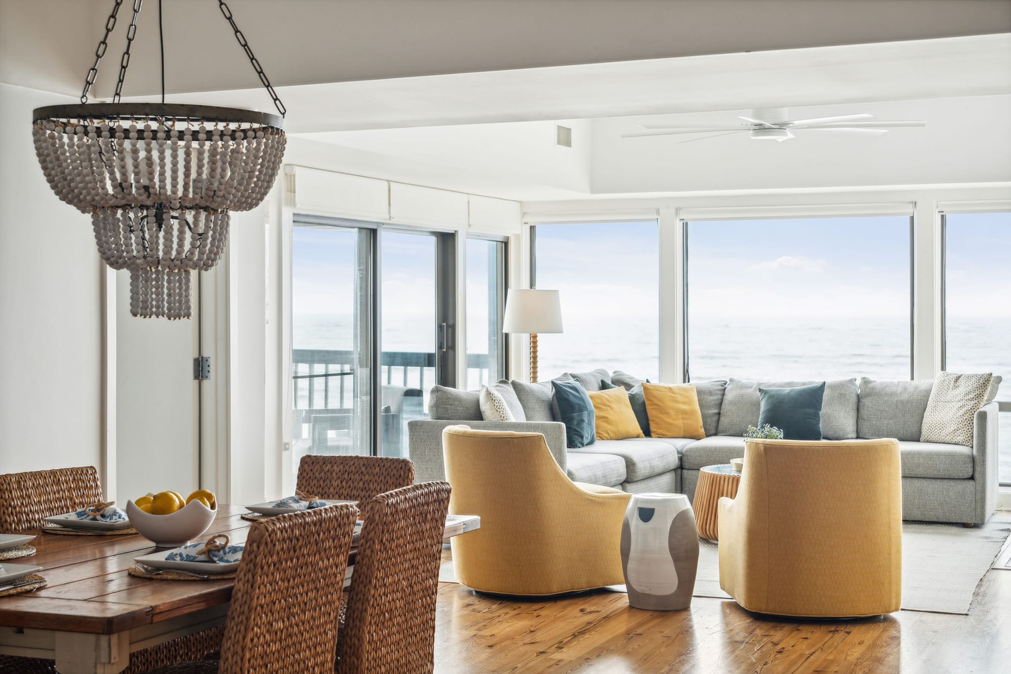 A bright, coastal living and dining area with floor-to-ceiling ocean views. The space features a cozy gray sectional sofa with mustard and navy pillows, woven dining chairs, a rustic wooden table set with blue and white plates, and a statement beaded chandelier. Sliding glass doors open to a balcony, enhancing the airy, beachside ambiance