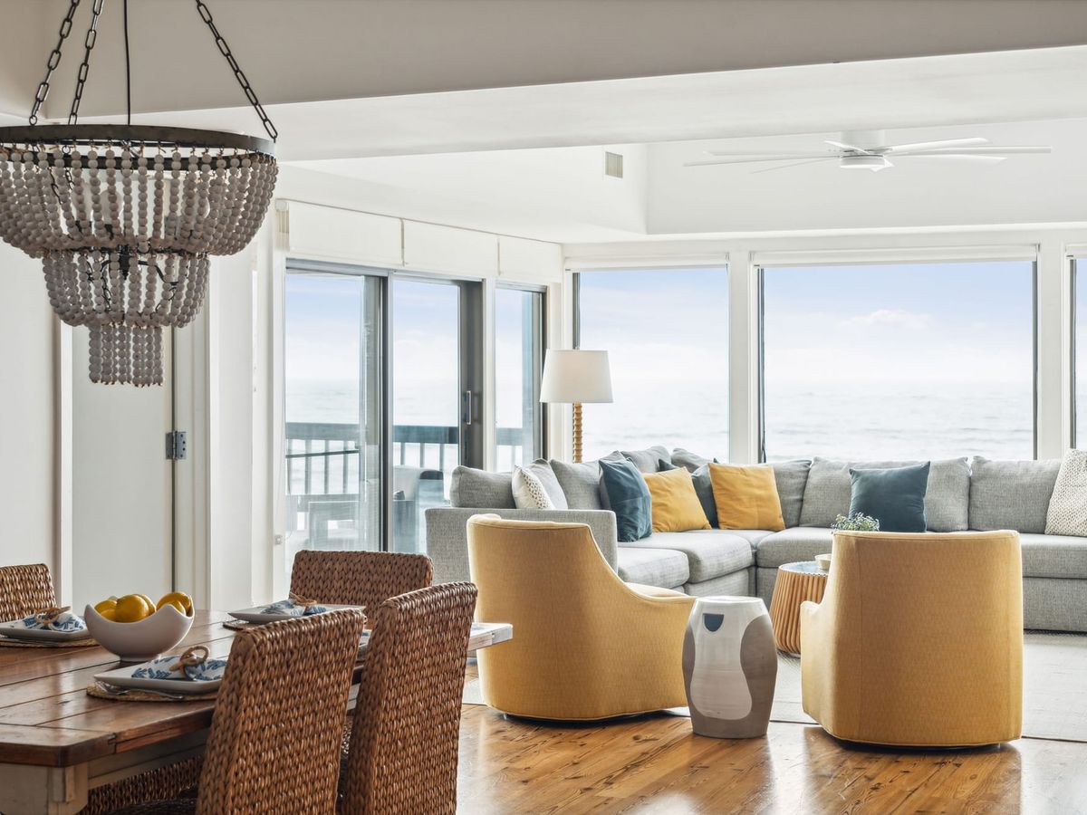 A bright, coastal living and dining area with floor-to-ceiling ocean views. The space features a cozy gray sectional sofa with mustard and navy pillows, woven dining chairs, a rustic wooden table set with blue and white plates, and a statement beaded chandelier. Sliding glass doors open to a balcony, enhancing the airy, beachside ambiance