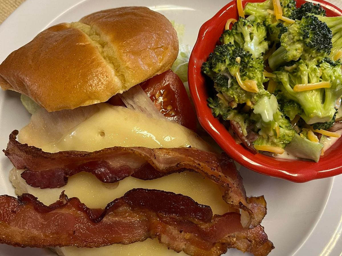 A bacon and cheese sandwich on a soft roll is plated with a fresh broccoli salad on the side. Country Cafe delivers homestyle comfort food in a simple and satisfying way.