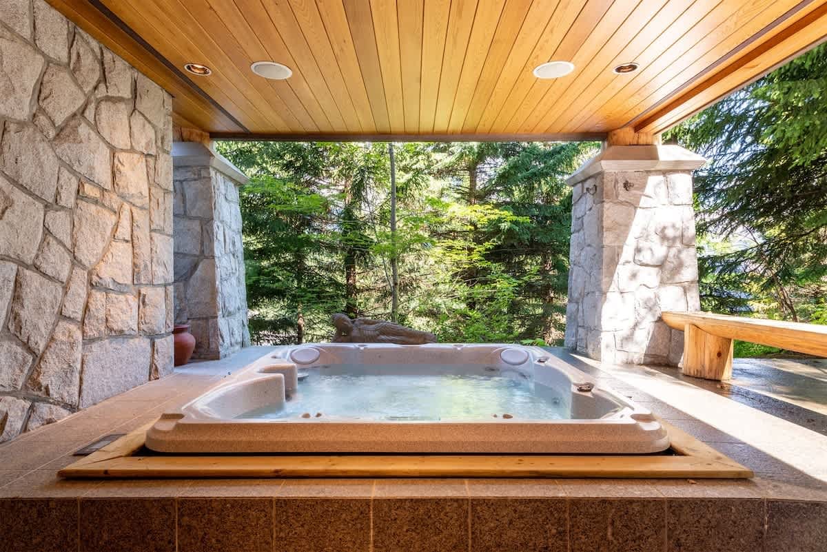 Covered outdoor hot tub set between stone pillars and a wood-paneled ceiling, overlooking a serene forest setting with lush green trees.