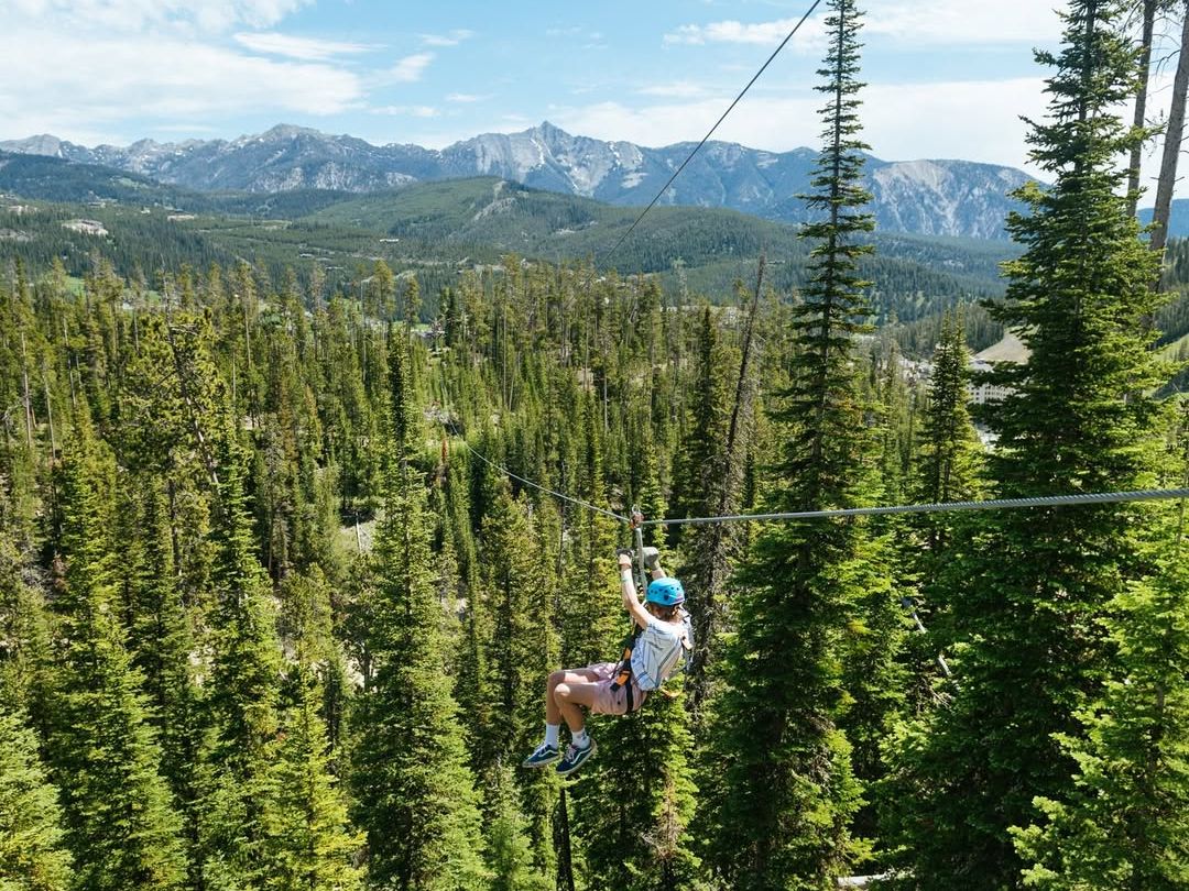 A person ziplines through a forest in Big Sky, Montana, with snow-capped mountains in the background. This thrilling ride gives you a bird’s-eye view of the trees and rugged peaks—perfect for summer adventure seekers.