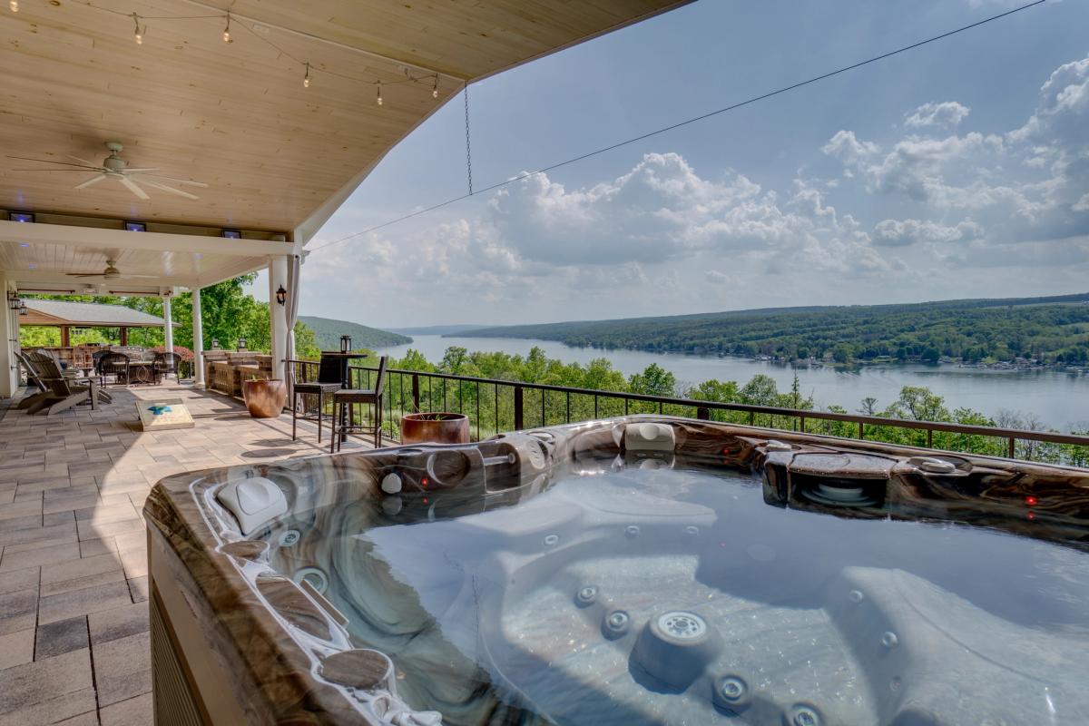 A luxurious outdoor patio with a hot tub overlooks a scenic lake surrounded by lush green hills. The covered seating area features lounge chairs, a bar-height table, and string lights, creating a relaxed and inviting atmosphere.