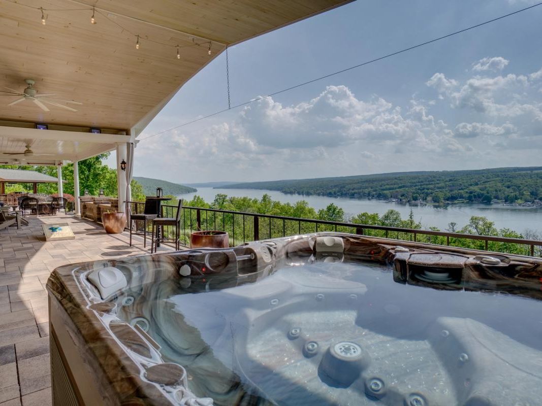A luxurious outdoor patio with a hot tub overlooks a scenic lake surrounded by lush green hills. The covered seating area features lounge chairs, a bar-height table, and string lights, creating a relaxed and inviting atmosphere.