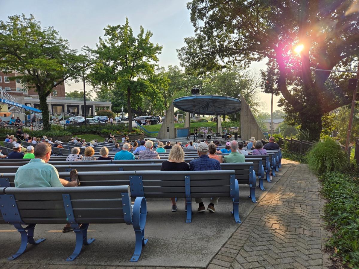 Visitors gather in an outdoor amphitheater as the sun sets behind tall trees, casting a warm golden light over the scene. The relaxed crowd enjoys live music in a charming small-town park setting.