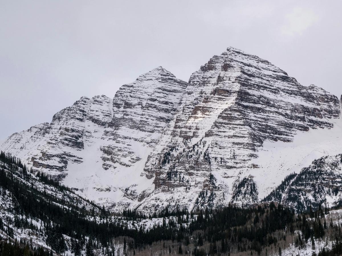 The iconic, snow-striped peaks of the Maroon Bells rise dramatically above a blanket of evergreens, standing stark and majestic beneath a wintry gray sky.