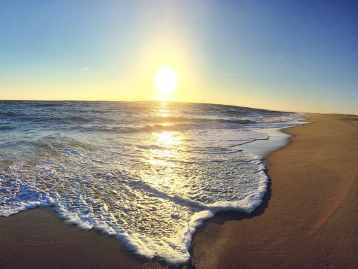 Golden sunset over a calm ocean, with gentle waves washing onto a wide, empty sandy beach under a clear sky.