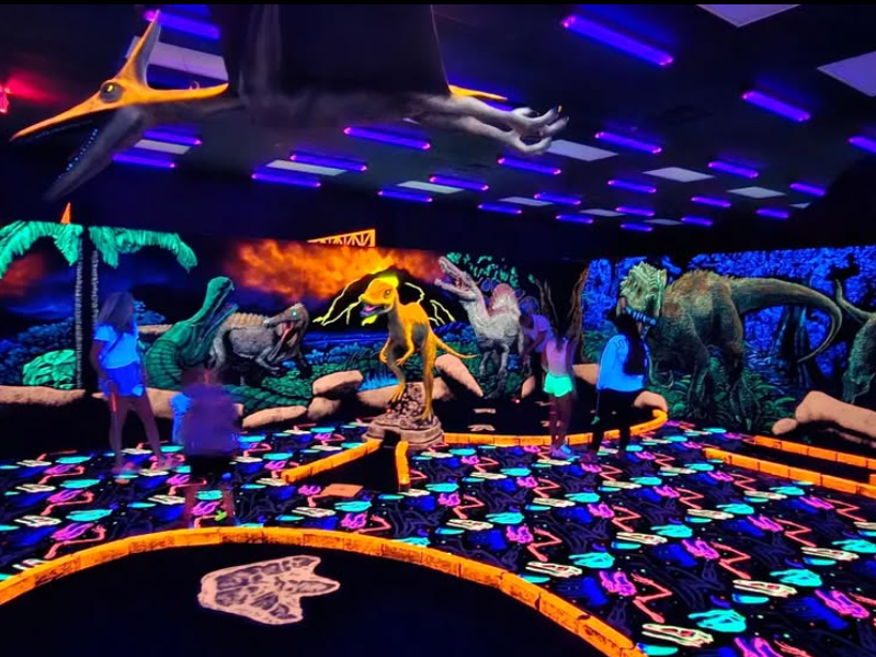 People play mini golf in a glowing, blacklight-illuminated course with vibrant dinosaur-themed decor and neon prehistoric scenery.