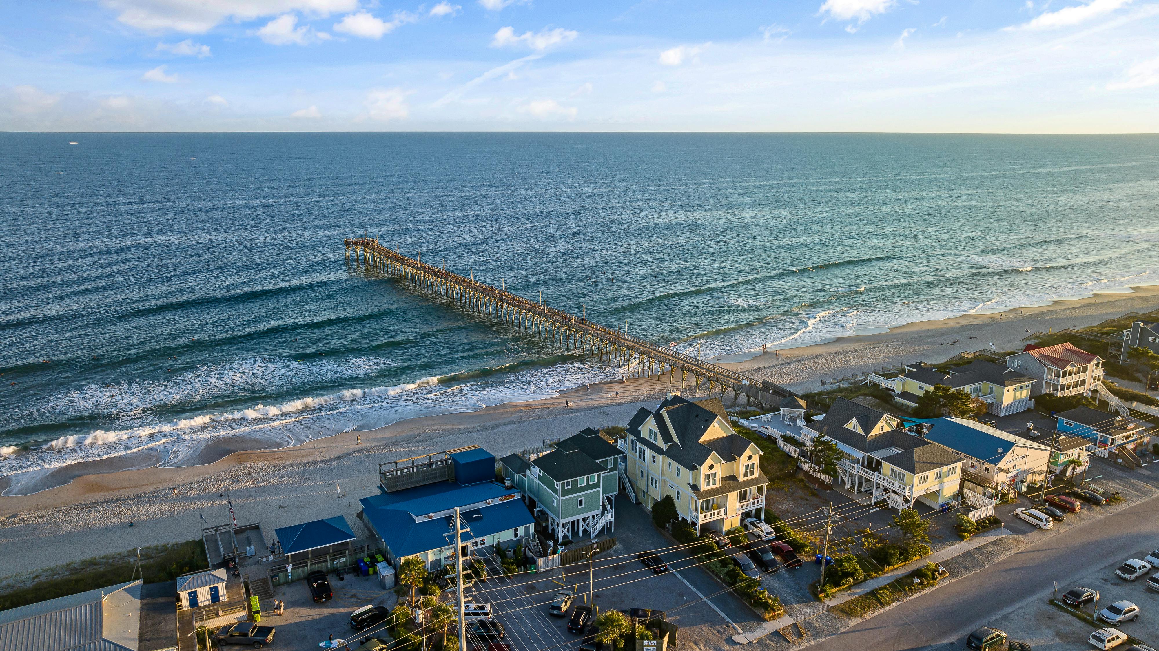 A stunning aerial view of a quiet beach town with a long fishing pier stretching into the ocean during golden hour. Soft waves roll onto the sandy shore while beachfront homes line the coast, creating the perfect North Carolina beach vacation scene.