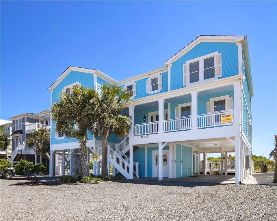 A bright blue, two-story beach house on stilts with white trim, a wraparound porch, and a covered parking area underneath. A staircase leads up to the main entrance, flanked by palm trees. The sky is clear and blue, and the gravel driveway extends to the front of the house. A rental sign is visible on the porch railing.