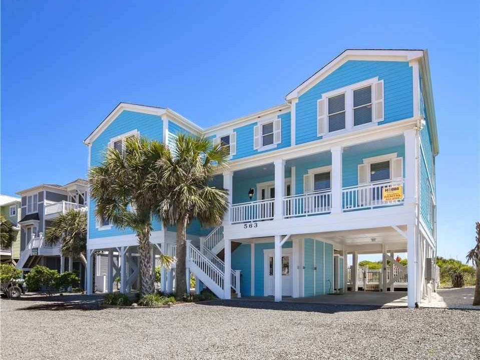 A bright blue, two-story beach house on stilts with white trim, a wraparound porch, and a covered parking area underneath. A staircase leads up to the main entrance, flanked by palm trees. The sky is clear and blue, and the gravel driveway extends to the front of the house. A rental sign is visible on the porch railing.