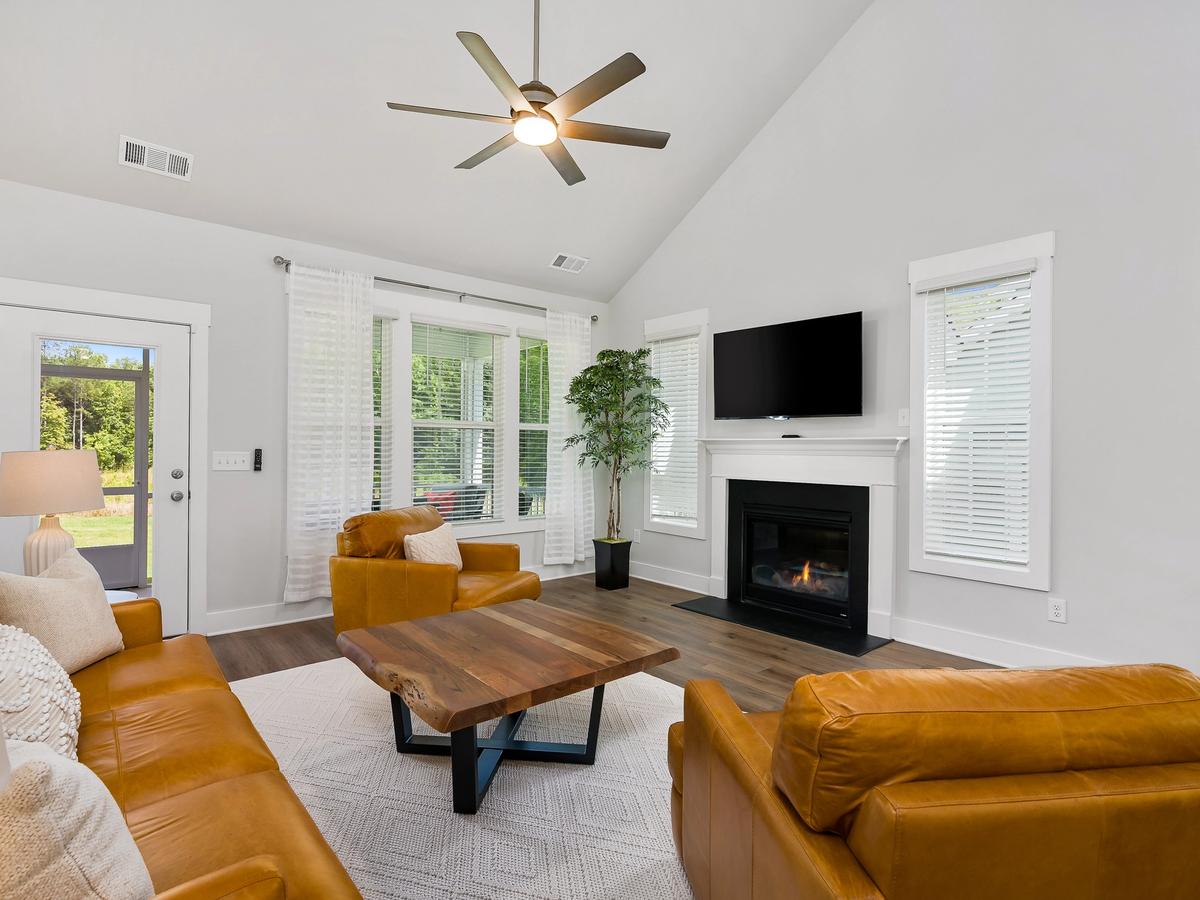 Bright living room with vaulted ceiling, tan leather sofa and chairs, a wooden coffee table, fireplace with mounted TV, large windows with white curtains, and a ceiling fan.