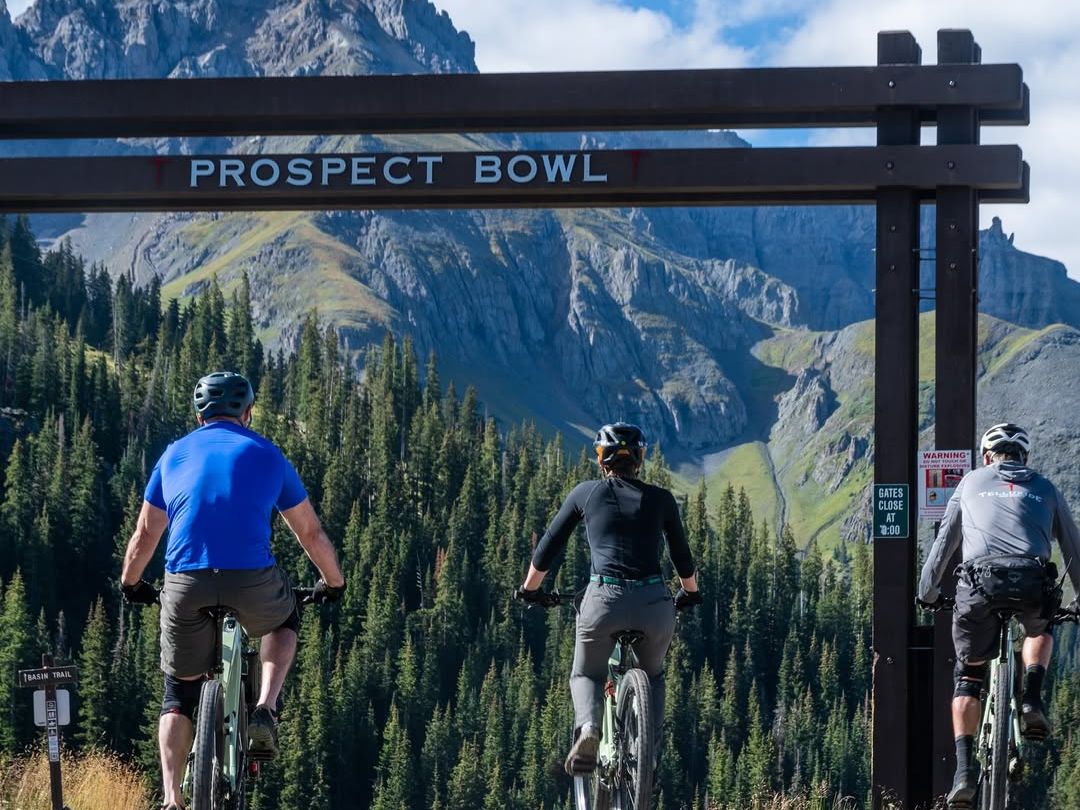 Three mountain bikers ride along Prospect Bowl trail under blue skies, with dramatic peaks and pine forests in the background. Summer in Telluride is perfect for hiking, biking, and outdoor fu