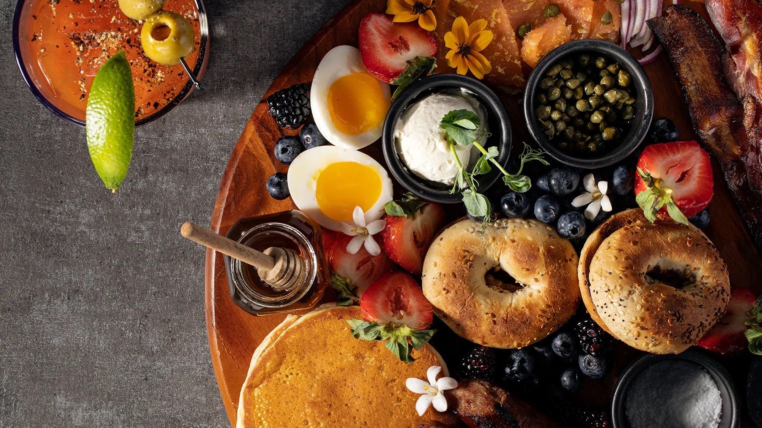 A cozy brunch spread featuring fresh bagels, eggs, berries, and pancakes sits beautifully arranged with honey and flowers. The rich colors and fresh ingredients invite you to slow down and savor every bite.