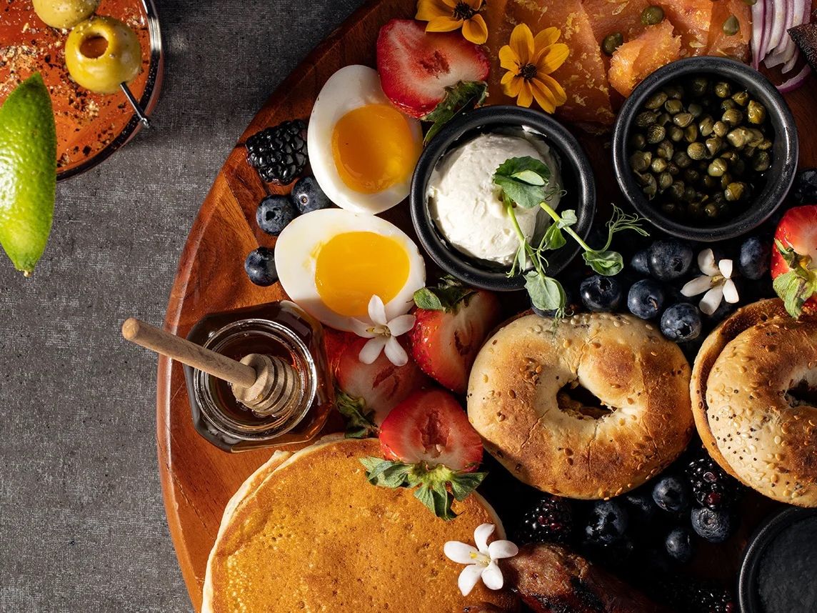 A cozy brunch spread featuring fresh bagels, eggs, berries, and pancakes sits beautifully arranged with honey and flowers. The rich colors and fresh ingredients invite you to slow down and savor every bite.