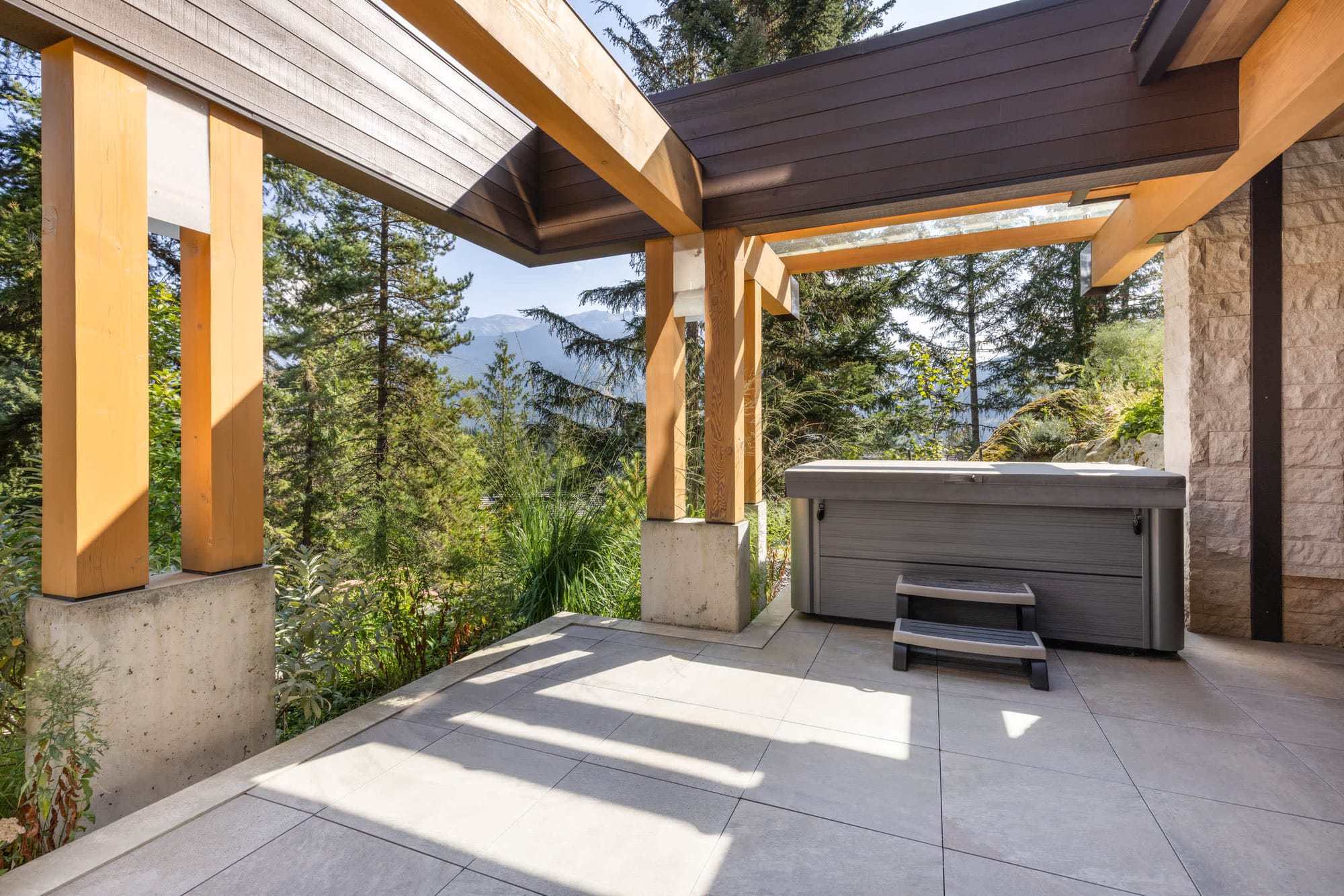 Covered mountain-view patio with exposed wood beams, stone accents, and a private hot tub overlooking lush forest and distant peaks.
