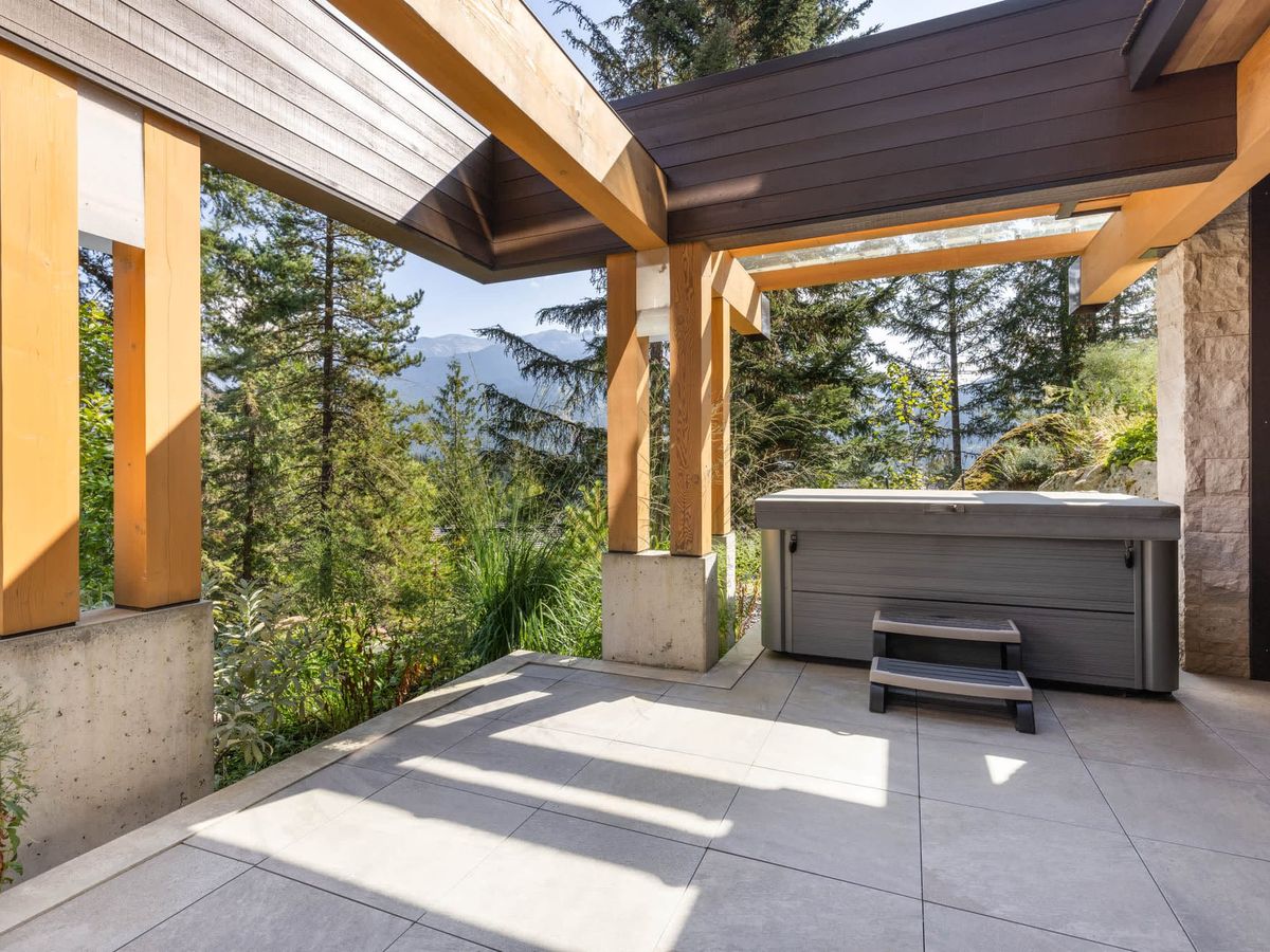 Covered mountain-view patio with exposed wood beams, stone accents, and a private hot tub overlooking lush forest and distant peaks.