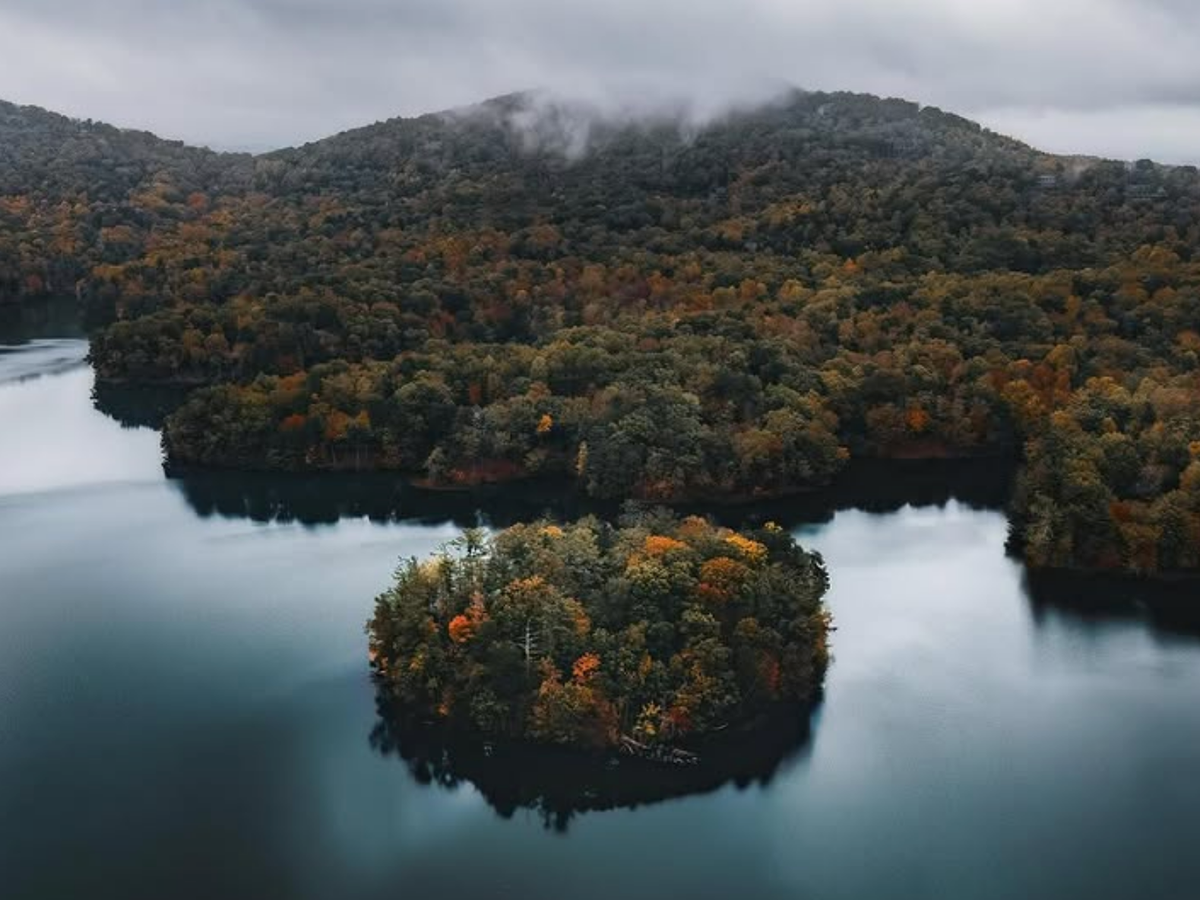 An aerial view of a misty mountain lake with dense autumn-colored forest and a small tree-covered island surrounded by calm, dark water.