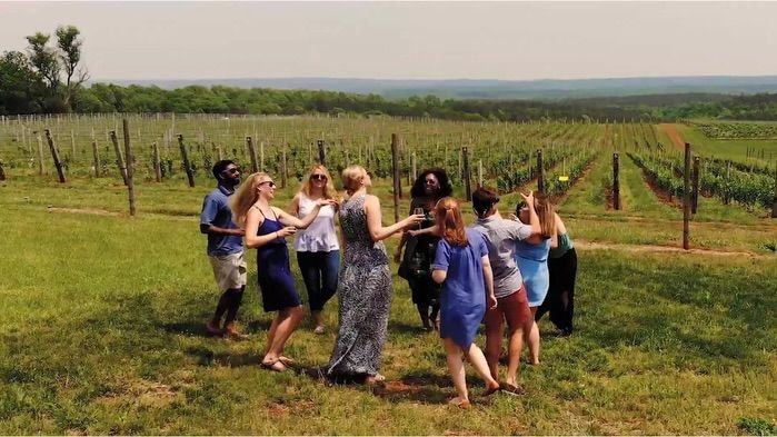 A group of friends gathers in the middle of a lush vineyard along the Monticello Wine Trail, laughing and raising glasses against a backdrop of endless vine rows. The rolling Virginia countryside stretches to the horizon under a hazy summer sky.