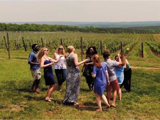 A group of friends gathers in the middle of a lush vineyard along the Monticello Wine Trail, laughing and raising glasses against a backdrop of endless vine rows. The rolling Virginia countryside stretches to the horizon under a hazy summer sky.