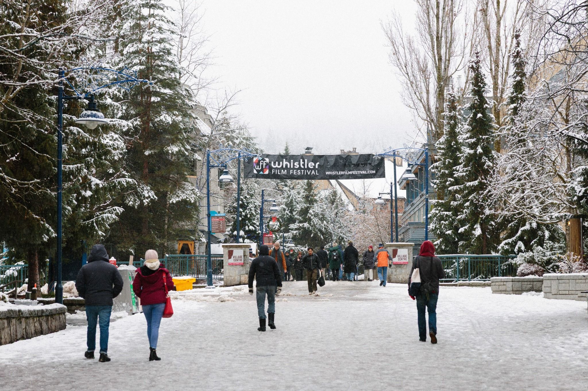 Visitors walk through snowy Whistler Village under a banner for the Whistler Film Festival. Snow-covered trees and winter decorations line the street as people explore the village. The festival brings film lovers together during the winter season in Whistler.