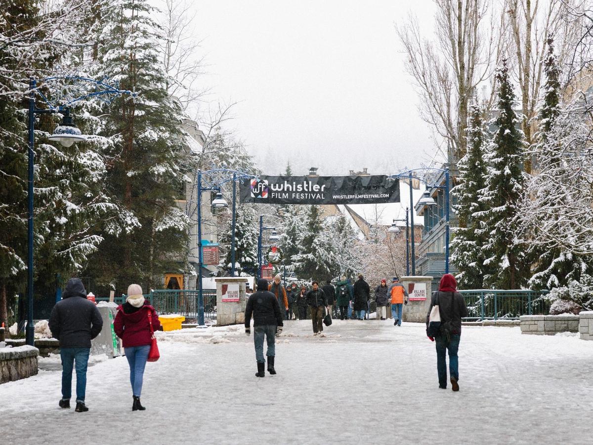 Visitors walk through snowy Whistler Village under a banner for the Whistler Film Festival. Snow-covered trees and winter decorations line the street as people explore the village. The festival brings film lovers together during the winter season in Whistler.