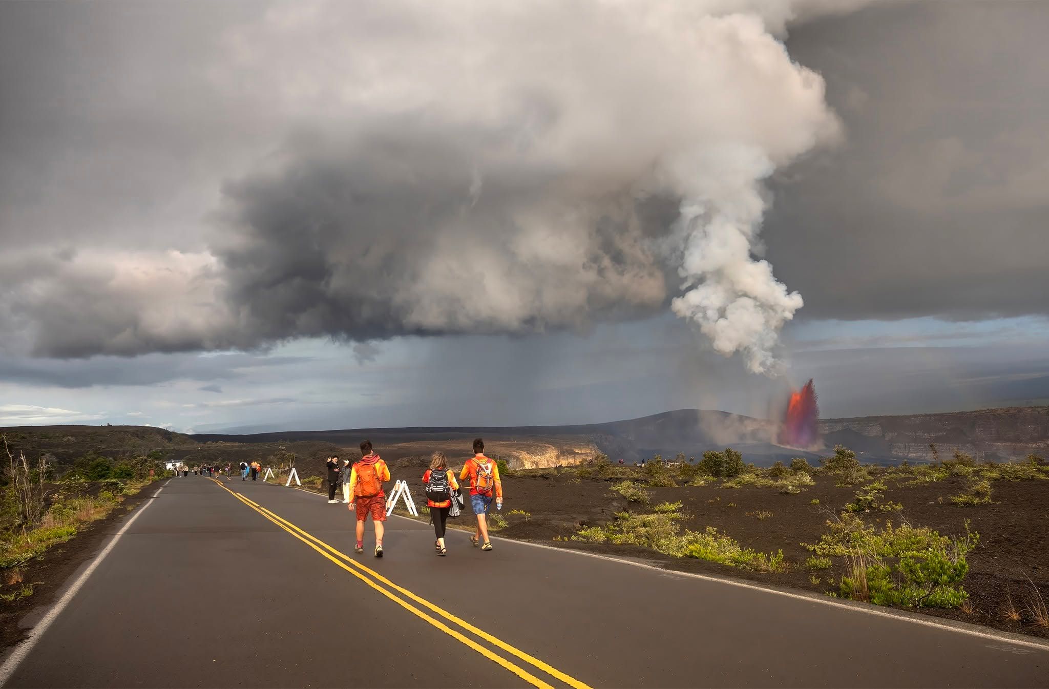 This shows Kīlauea, one of the world's most active volcanoes. You can see a lava fountain erupting on the right, sending up a tall plume of volcanic gas and steam, while visitors walk along a park road with the caldera and lava fields stretching out. The dramatic mix of storm clouds and eruption is characteristic of the park during active eruption periods.