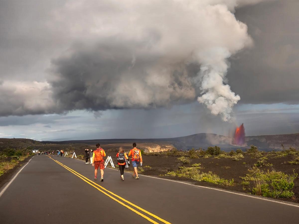 This shows Kīlauea, one of the world's most active volcanoes. You can see a lava fountain erupting on the right, sending up a tall plume of volcanic gas and steam, while visitors walk along a park road with the caldera and lava fields stretching out. The dramatic mix of storm clouds and eruption is characteristic of the park during active eruption periods.