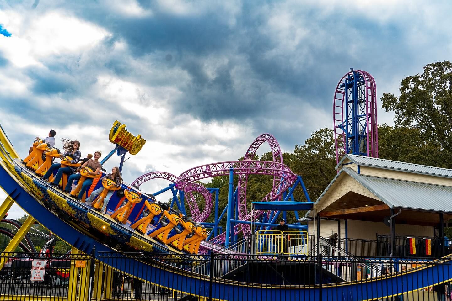 Riders enjoy a colorful spinning attraction at Oaks Amusement Park with twisting roller-coaster tracks behind them. The bold blue and yellow ride stands out against the cloudy sky. It captures the fun and excitement of a family day at the park.