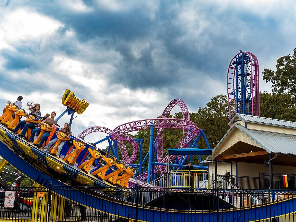 Riders enjoy a colorful spinning attraction at Oaks Amusement Park with twisting roller-coaster tracks behind them. The bold blue and yellow ride stands out against the cloudy sky. It captures the fun and excitement of a family day at the park.