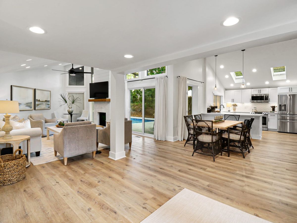 A super stylish, fully renovated coastal home features a bright open-plan living area with modern white sofas, crisp kitchen finishes, and a private pool with custom evening lightscaping, set against serene lagoon and golf views