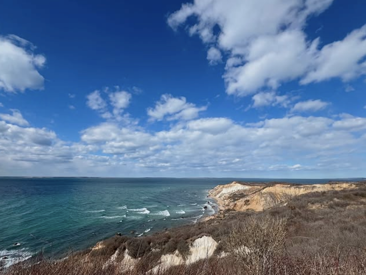 Scenic view of rugged coastal cliffs and turquoise ocean waves under a bright blue sky filled with scattered white clouds