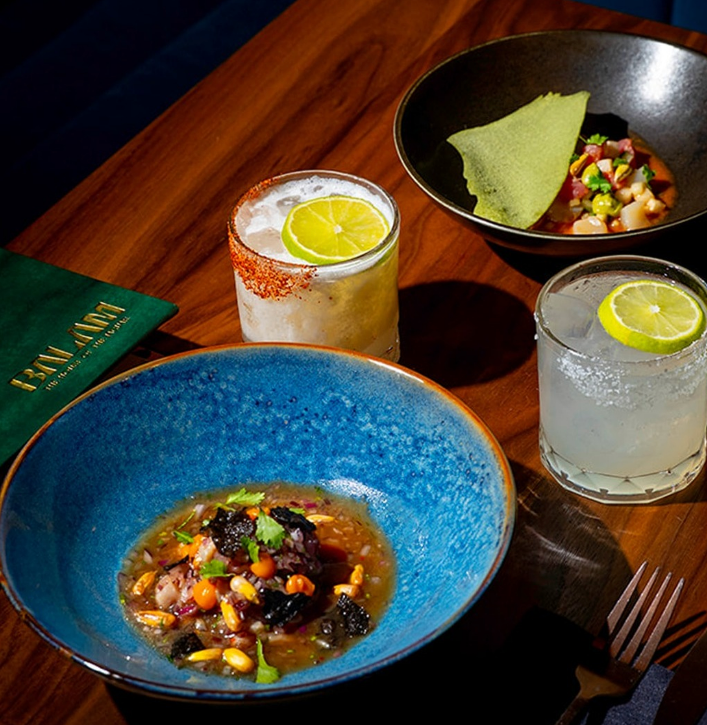 Vibrant small plates of ceviche served in colorful bowls alongside salt-rimmed margaritas garnished with lime, set on a warm wooden table in a stylish dining setting.