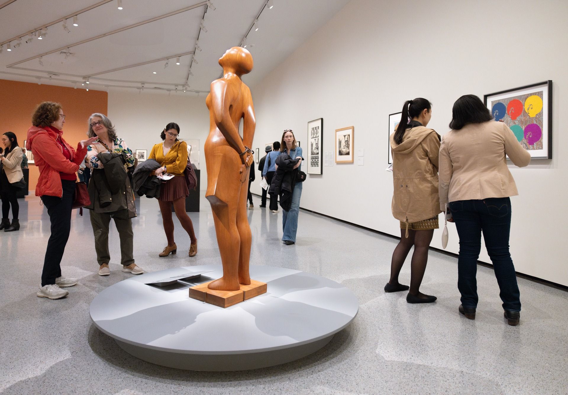Visitors move slowly through a bright, open gallery filled with sculptures and colorful framed artwork. A tall wooden sculpture stands in the center, catching the eye as people chat and study the art around them. The space feels lively and inspiring, perfect for an afternoon exploring culture in D.C.