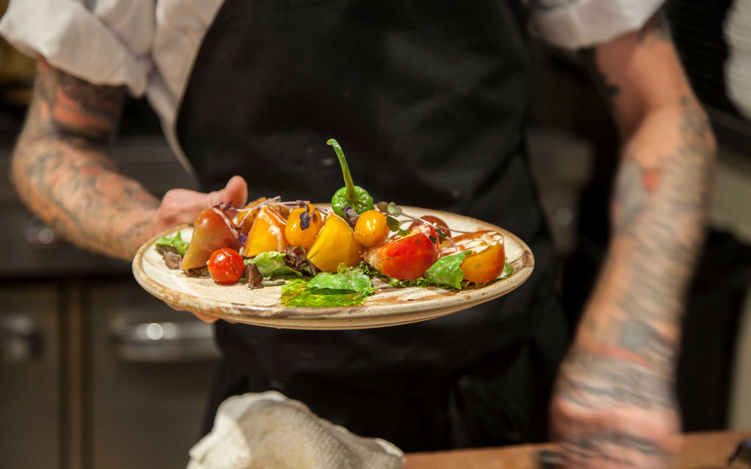 A chef holds out a beautifully plated dish filled with colorful tomatoes, fresh greens, and bright garnishes. The vibrant ingredients pop against the rustic plate, showing off the restaurant’s creative, farm-fresh style. The tattooed arms of the chef add a personal, artistic touch to the moment.
