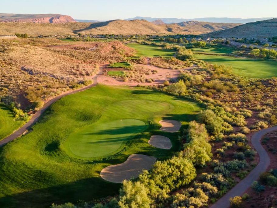 A stunning aerial view of a golf course set in a desert landscape, featuring lush green fairways, sand bunkers, and rolling hills with a backdrop of red rock formations and distant mountains