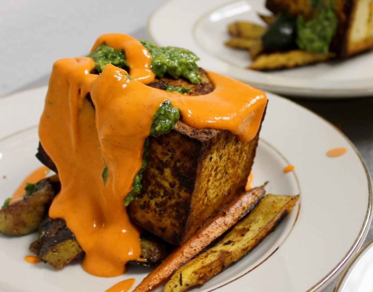 gourmet dish featuring a cube of toasted bread topped with green herb pesto and draped in creamy orange sauce, served with roasted vegetables.