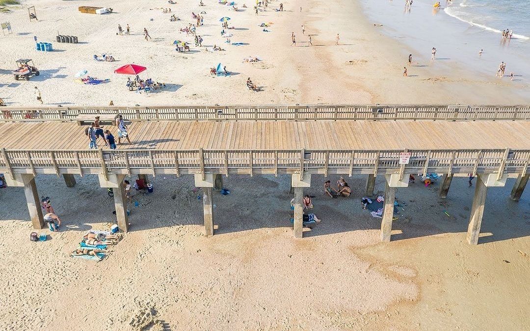 An aerial view of Tybee Pier and Pavilion shows beachgoers relaxing under umbrellas and strolling along the sandy shore. The lively scene captures the heart of Tybee Island’s beach culture.