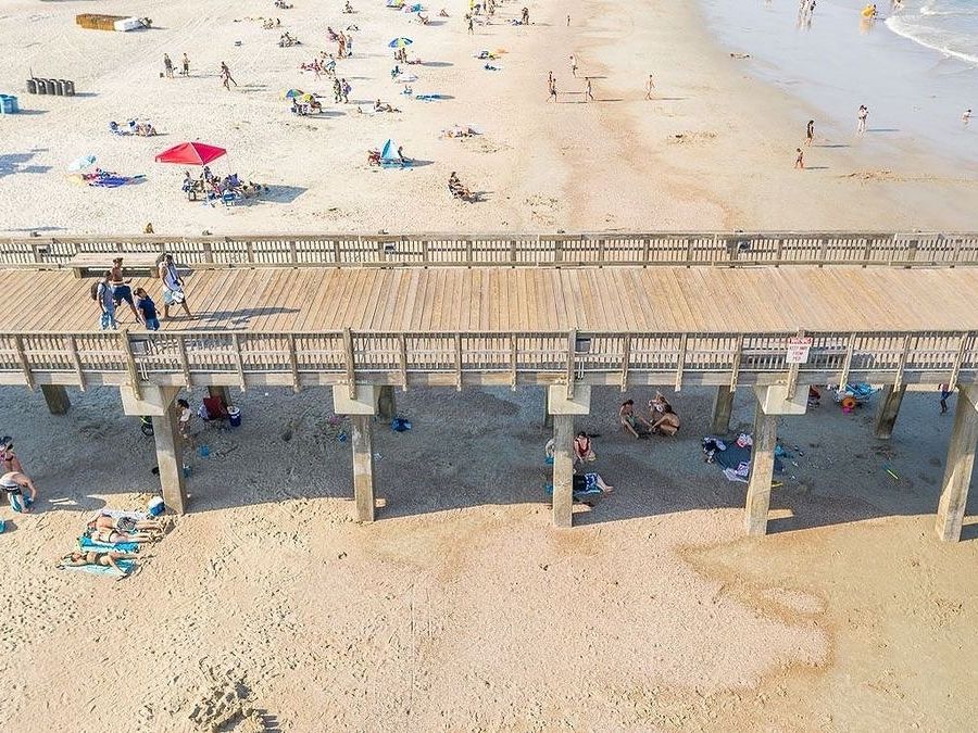 An aerial view of Tybee Pier and Pavilion shows beachgoers relaxing under umbrellas and strolling along the sandy shore. The lively scene captures the heart of Tybee Island’s beach culture.