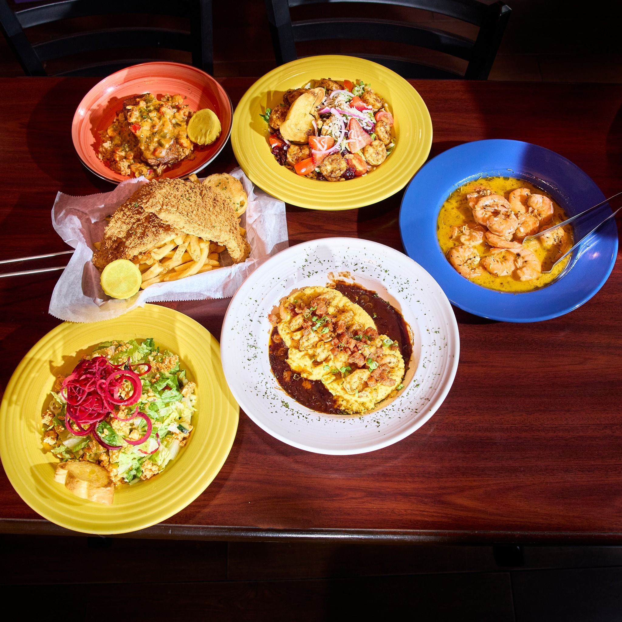 A colorful table filled with New Orleans-style dishes like fried seafood, creamy pastas, and bright salads creates a lively, comfort-food spread. The bold colors and generous portions make everything look rich and full of flavor. It feels like a fun, hearty meal you’d enjoy with a big group.