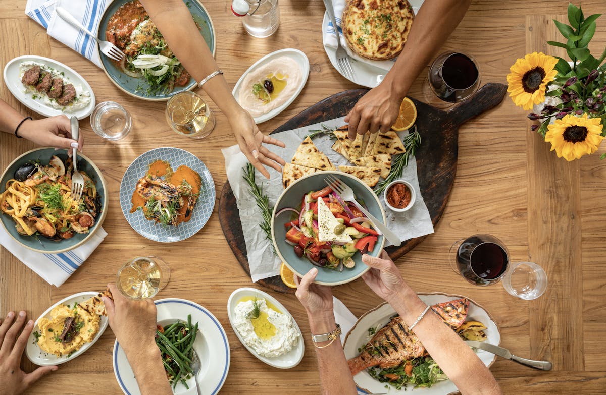 A table filled with Greek dishes like fresh salad, grilled seafood, and warm pita at Kipos Hellenic Cuisine. The family-style spread, shared with wine and friends, celebrates Mediterranean flavors and togetherness.