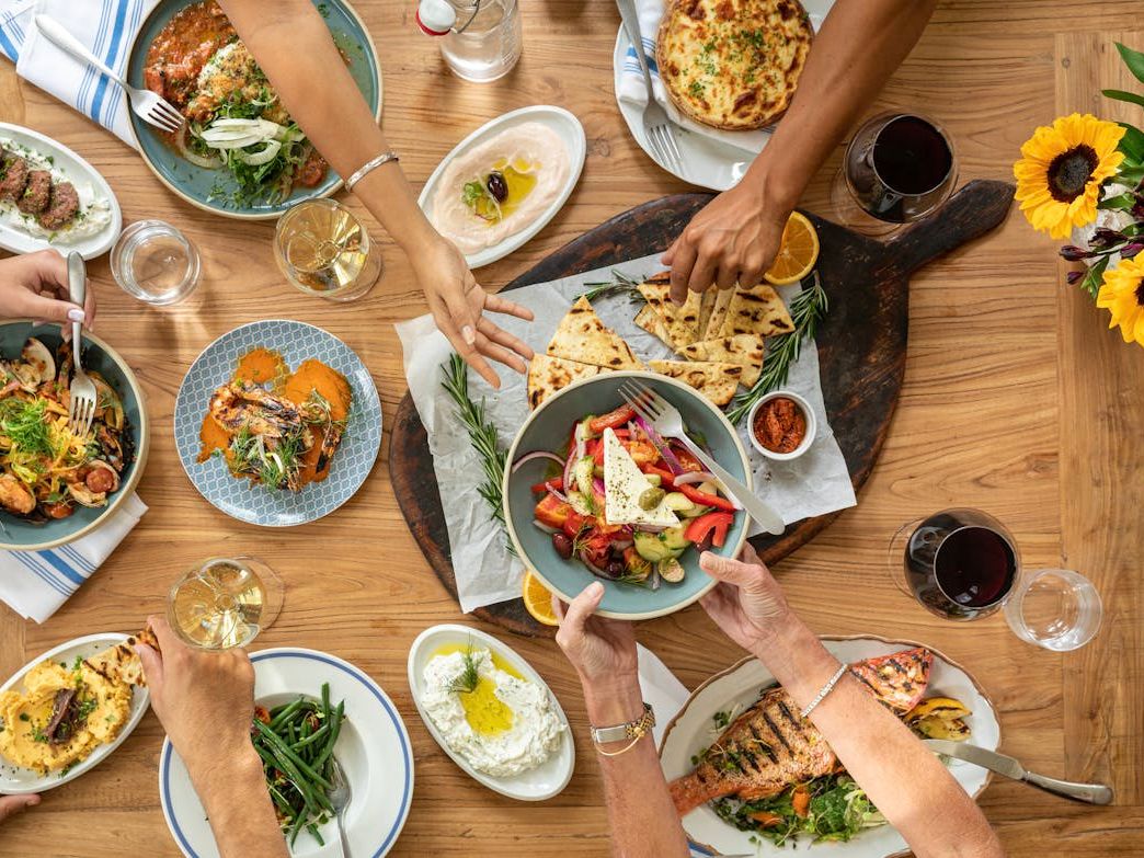 A table filled with Greek dishes like fresh salad, grilled seafood, and warm pita at Kipos Hellenic Cuisine. The family-style spread, shared with wine and friends, celebrates Mediterranean flavors and togetherness.