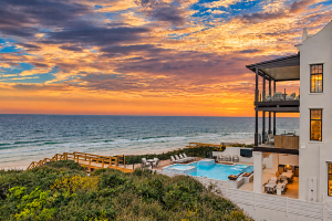 A white beachfront home overlooks the ocean beneath a soft pink and blue evening sky. The outdoor area includes a pool, lounge chairs, covered balconies, and wooden stairs leading toward the beach.