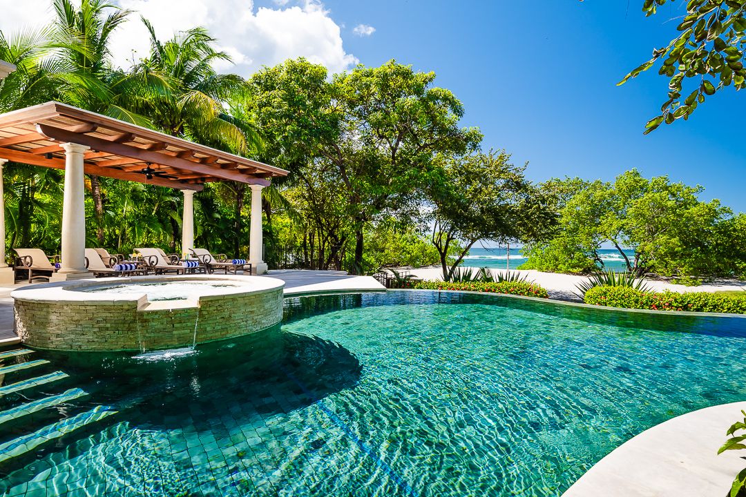 A luxurious beachfront pool surrounded by lush greenery, with cushioned loungers under a shaded pergola and a glimpse of the ocean just beyond the trees.