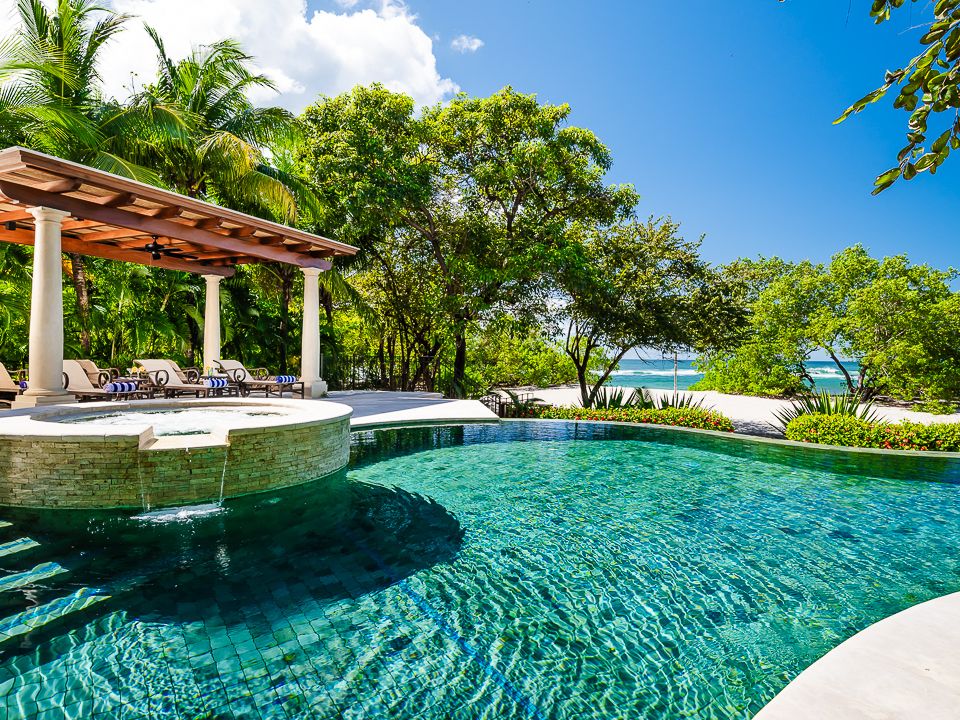 A luxurious beachfront pool surrounded by lush greenery, with cushioned loungers under a shaded pergola and a glimpse of the ocean just beyond the trees.