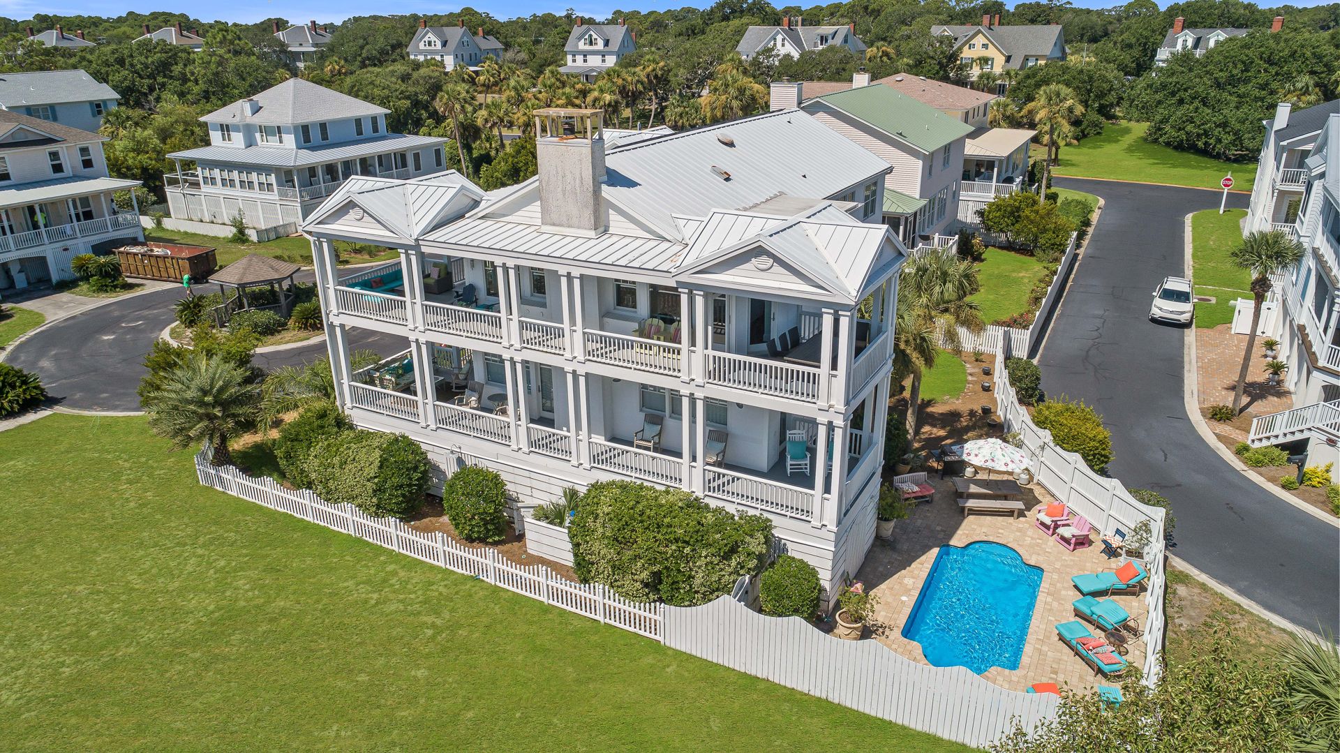 Aerial view of a large white coastal home with multiple wraparound porches and a private fenced-in backyard featuring a pool, colorful lounge chairs, and umbrella seating, set in a quiet beachside neighborhood.