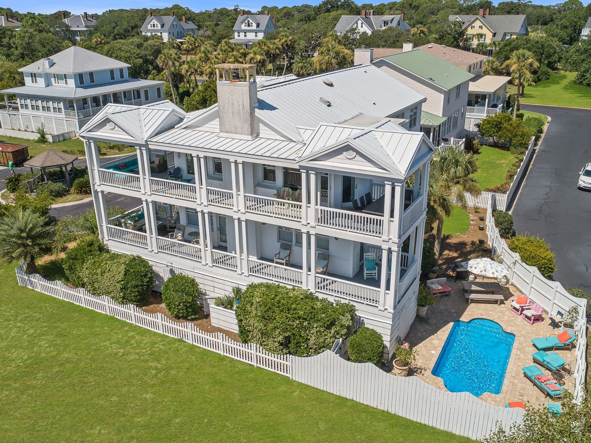 Aerial view of a large white coastal home with multiple wraparound porches and a private fenced-in backyard featuring a pool, colorful lounge chairs, and umbrella seating, set in a quiet beachside neighborhood.