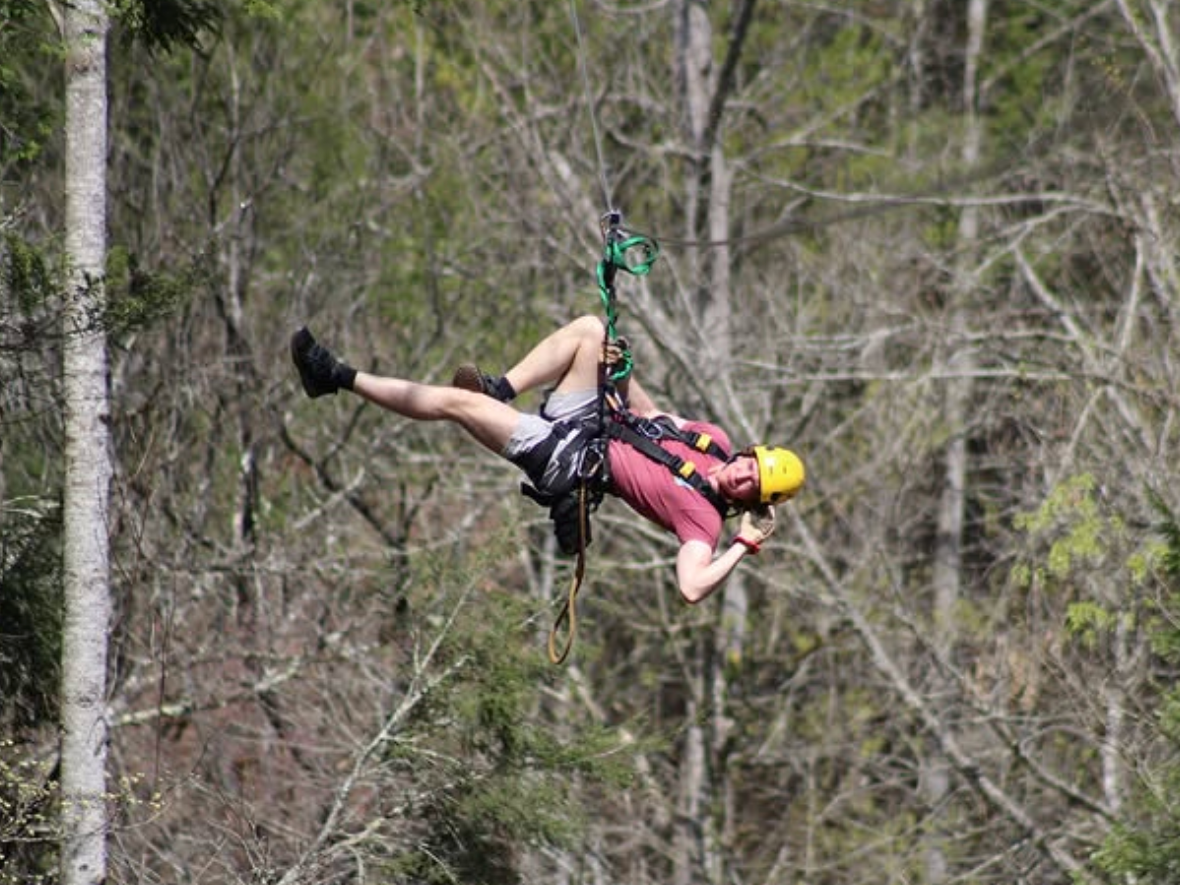 A person wearing a helmet and harness hangs playfully sideways while ziplining through a forest.