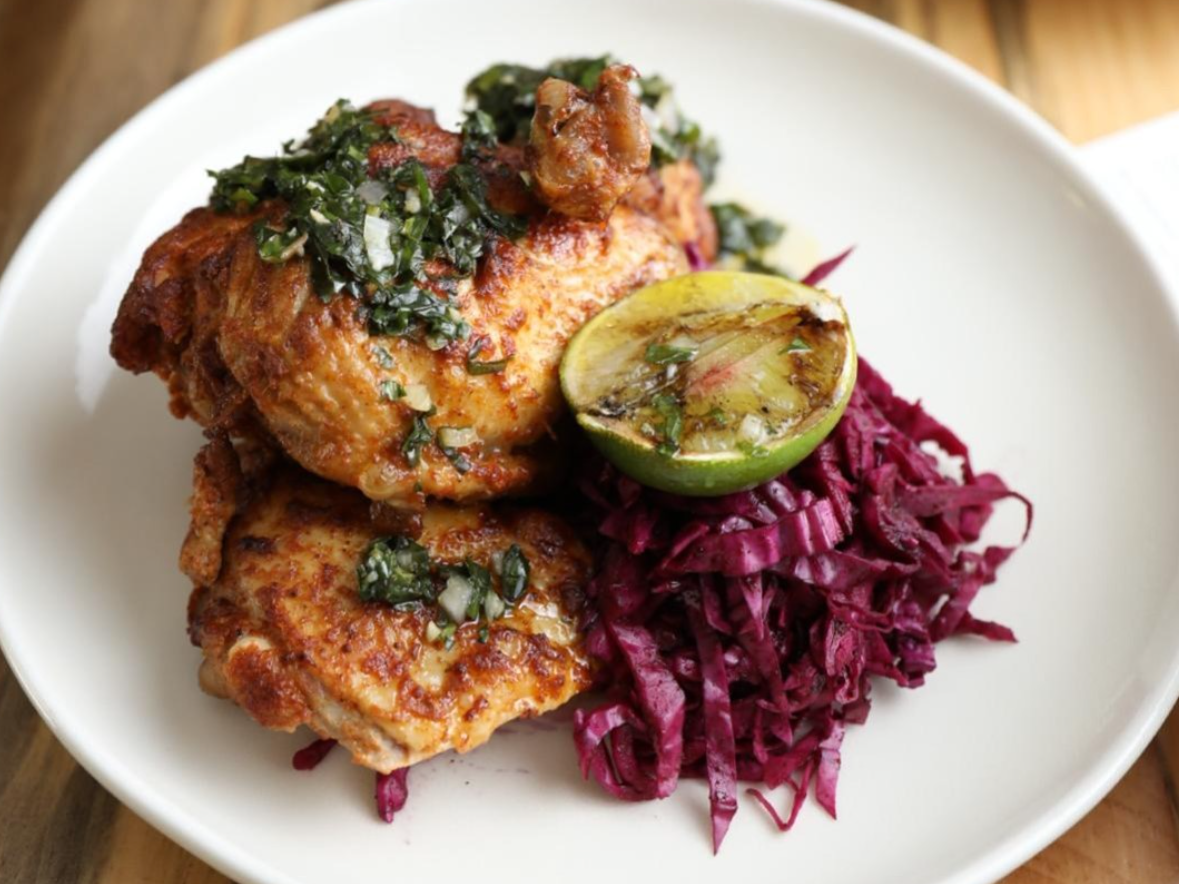 A plate of grilled chicken thighs topped with herb sauce is served alongside tangy red cabbage slaw and a charred lime half.