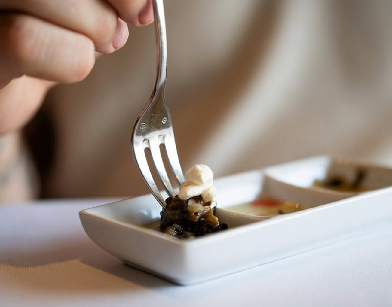 A close-up of a fork lifting a small bite of gourmet food topped with cream from a white tasting dish.