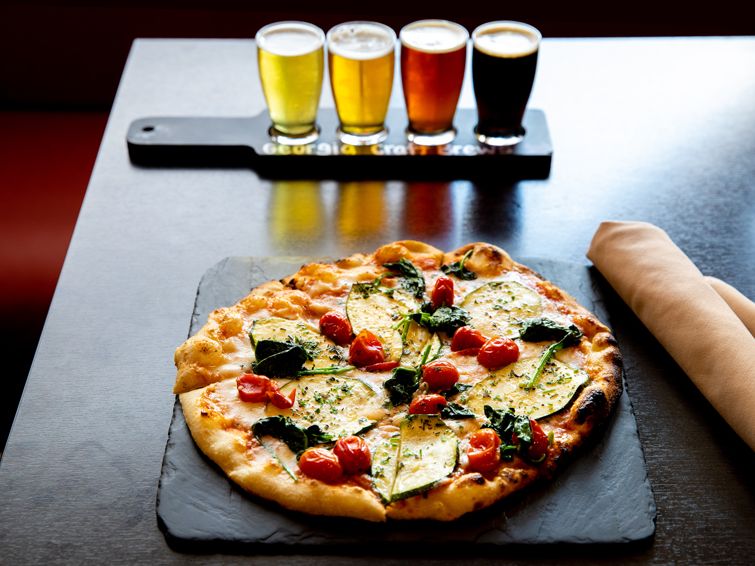 A wood-fired veggie pizza topped with cherry tomatoes, zucchini, and greens sits next to a colorful flight of craft beers at The Nest. Great for casual nights and local flavor lovers.